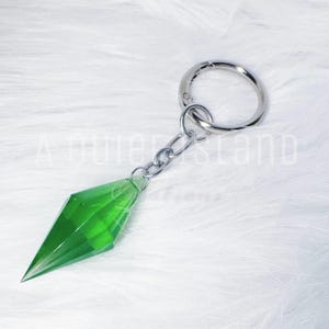 May include: A keychain featuring a translucent, emerald green, diamond-shaped pendant with faceted edges. The pendant is attached to a silver-toned metal jump ring and a circular split ring, providing a secure attachment point for keys or accessories.