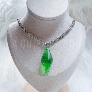 May include: A necklace featuring a translucent, bright green, diamond-shaped pendant suspended from a silver-toned metal chain. The pendant has a faceted, geometric design that tapers to a point at the bottom. The text A QUIET LAND is visible.
