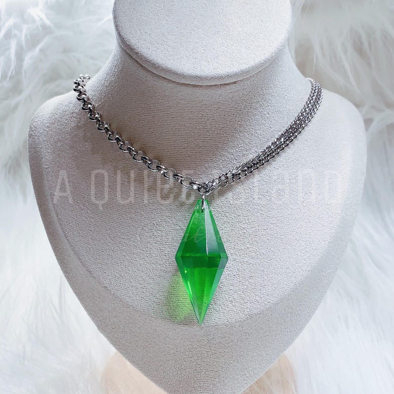 May include: A necklace featuring a translucent, bright green, diamond-shaped pendant suspended from a silver-toned metal chain. The pendant has a faceted, geometric design that tapers to a point at the bottom. The text A QUIET LAND is visible.