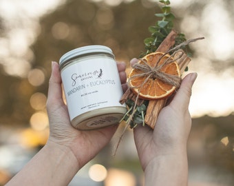 May include: A white candle jar with a white label that reads "Somerville Gardens" and "Mandarin + Eucalyptus" with a 9 fl oz soy candle description. The candle is being held in a person's hand along with a bundle of dried orange slices, cinnamon sticks, and eucalyptus.