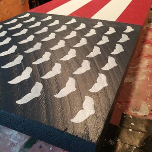 May include: A wooden sign painted with an American flag design. The sign features a blue field with white stars and red and white stripes.
