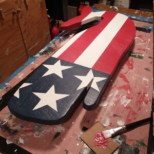 May include: A wooden hand-shaped sign painted in the colors of the American flag. The sign is painted red, white, and blue with white stars on a blue background. A small paintbrush and a dab of red paint are visible in the foreground.