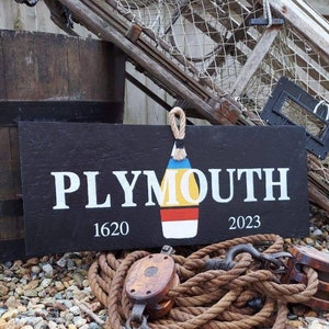 May include: Black wooden sign with white text that reads "PLYMOUTH 1620 2023". A colorful buoy is painted in the center of the sign.