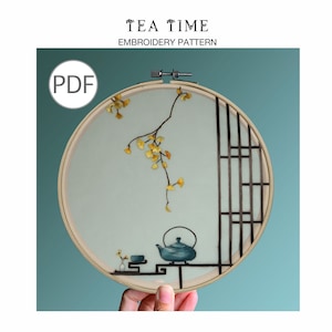 Könnte beinhalten: Stickmuster-PDF für eine Teestunde-Szene. Das Muster zeigt eine Teekanne, eine Teetasse und einen Zweig mit gelben Blättern in einem hellblauen Reifen.