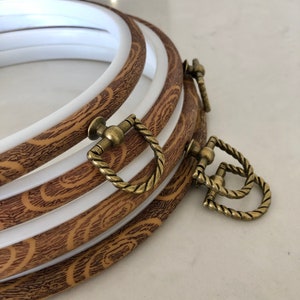 Peut inclure: Ensemble de quatre cerceaux à broder avec un motif en bois brun et une bordure blanche. Les cerceaux ont des fermoirs de couleur laiton.