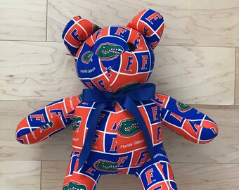 May include: A blue and orange teddy bear with a Florida Gators pattern. The bear has a blue bow tie and is sitting on a wooden surface.