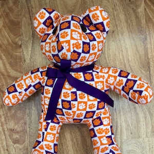 May include: A handmade teddy bear with a checkered pattern of orange and purple paw prints on a white background. The bear has a purple ribbon tied around its neck. The bear is a plush toy.