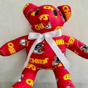 May include: A red and yellow teddy bear with a white bow. The bear is covered in the Kansas City Chiefs logo and the words "Chiefs" are printed on the fabric.