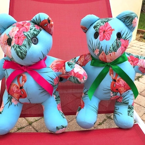 May include: Two plush teddy bears with light blue bodies and floral patterned fabric on their heads, arms, and legs. The floral pattern includes pink hibiscus flowers and red flamingos. One bear has a pink bow, the other a green bow.