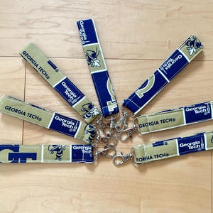 May include: A collection of fabric key fobs featuring the Georgia Tech logo and colors. The fobs are in gold, navy blue, and white, with the words "GEORGIA TECH" printed on them. Each fob has a silver-colored metal clasp.