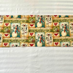 May include: A rectangular fabric table runner featuring a repeating pattern of snowmen, red birds, and hearts on a cream and green background. The design includes the text "WINTER WONDERS" printed in a decorative font across the horizontal sections.