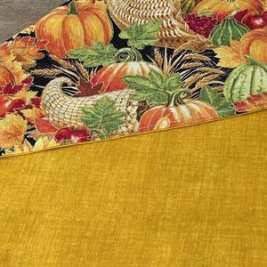 May include: A fall-themed table runner featuring a black background with pumpkins, sunflowers, and cornucopias. The bottom portion is a solid mustard yellow. The runner is likely made of fabric and suitable for seasonal home decor.
