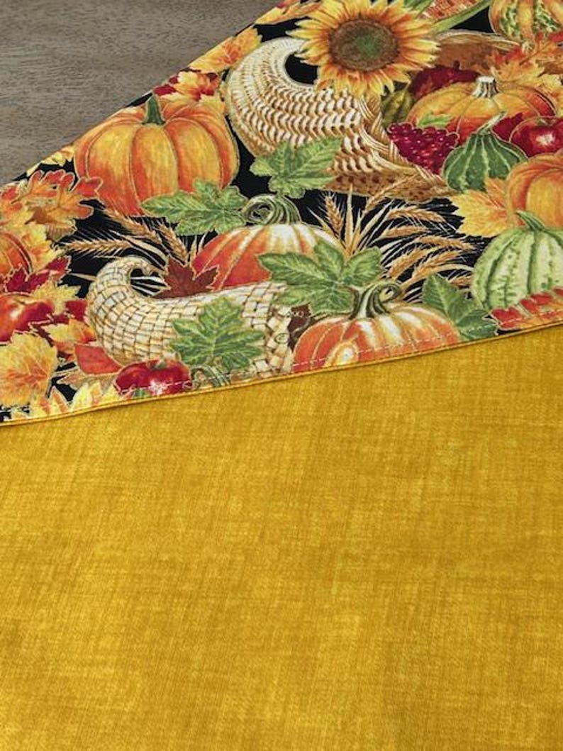 May include: A fall-themed table runner featuring a black background with pumpkins, sunflowers, and cornucopias. The bottom portion is a solid mustard yellow. The runner is likely made of fabric and suitable for seasonal home decor.