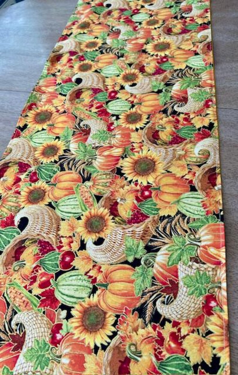 May include: A rectangular table runner with a colorful autumn harvest pattern. The design features sunflowers, pumpkins, gourds, cornucopias, and fall leaves in shades of orange, yellow, red, and green on a black background.