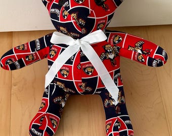 May include: A handmade teddy bear featuring a repeating pattern of the Florida Panthers logo in red, navy blue, and white. The bear has a white ribbon tied around its neck. The bear is made of fabric and has a soft, plush appearance.