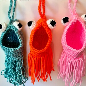 Könnte beinhalten: Drei gehäkelte Hängeschmuckstücke in Form von Fischen mit großen Glitz Augen. Die Fische sind türkis, orange und rosa.