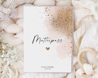 May include: A white card with a pink and gold watercolor background. The text "Mutterpass" is written in black script with a pink heart below it. The name "JULIA FRIEBE" and the date "03.06.90" are printed below the heart.