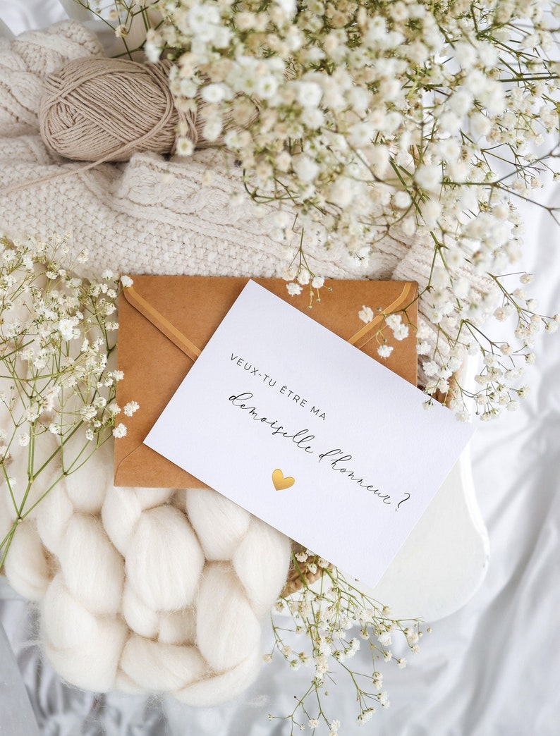 May include: A white card with the text "veux-tu etre ma demoiselle d'honneur?" and a gold heart. The card is on a brown envelope and surrounded by white flowers.