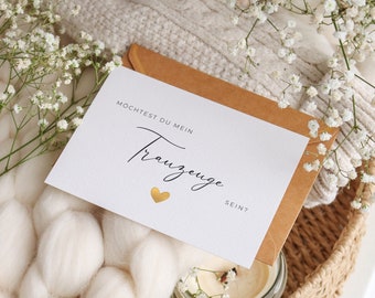 May include: A white card with gold lettering that says "Mochtest du mein Trauzeuge sein?" with a gold heart. The card is on a brown envelope and surrounded by white flowers and a white fluffy blanket.