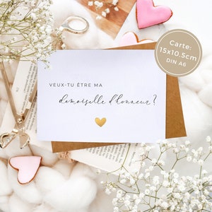 May include: A white card with the text "veux-tu &ecirc;tre ma demoiselle d'honneur?" in black script. A small gold heart is below the text. The card is on a brown surface with white flowers.