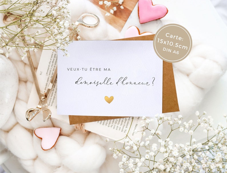 May include: A white card with the text "veux-tu &ecirc;tre ma demoiselle d'honneur?" in black script. A small gold heart is below the text. The card is on a brown surface with white flowers.