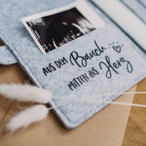 May include: A light blue felt photo album with a snap closure. The album holds a black and white ultrasound image and has the German text "AUS DEM BAUCH... MITTEN INS HERZ" in black script. A sprig of white dried flowers rests in front.