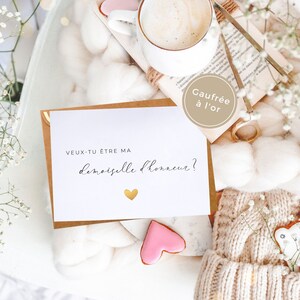 May include: A white card with the text "Veux-tu &ecirc;tre ma demoiselle d'honneur?" in black script with a gold heart. The card is on a white fuzzy surface with a brown envelope, a book, a cup of coffee, and pink and white cookies.