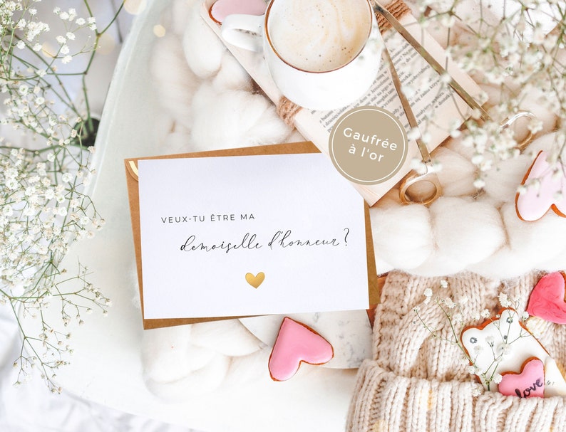 May include: A white card with the text "Veux-tu &ecirc;tre ma demoiselle d'honneur?" in black script with a gold heart. The card is on a white fuzzy surface with a brown envelope, a book, a cup of coffee, and pink and white cookies.
