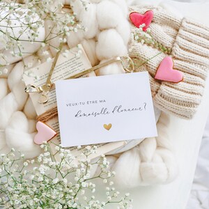 May include: A white card with the text "VEUX-TU &Ecirc;TRE MA DEMOISELLE D'HONNEUR?" in black lettering and a gold heart. The card is surrounded by white flowers, a white knitted hat, and a book.
