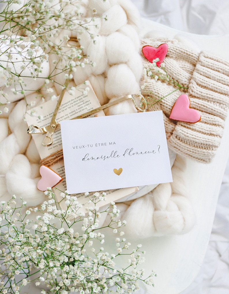 May include: A white card with the text "VEUX-TU &Ecirc;TRE MA DEMOISELLE D'HONNEUR?" in black lettering and a gold heart. The card is surrounded by white flowers, a white knitted hat, and a book.