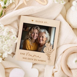 Pode incluir: Uma moldura de madeira clara com uma fotografia de duas mulheres sorrindo. A moldura tem o texto alemão "Glück ist... Eine Mama wie dich zu haben!" e um design floral.