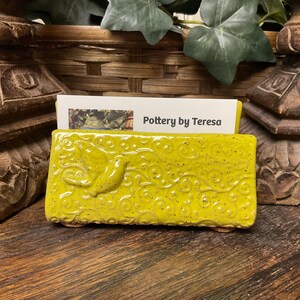 May include: A rectangular, yellow ceramic business card holder with a bird and swirl design. The holder has a speckled finish and the text "Pottery by Teresa" is visible on a card inside.