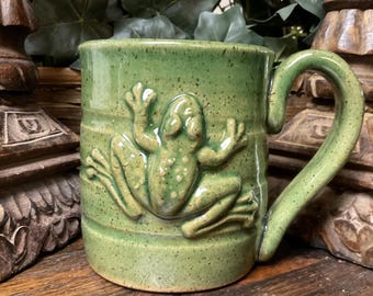 May include: A green ceramic mug with a speckled finish and a raised, three-dimensional frog relief on the front. The mug features a sturdy, curved handle and a cylindrical body, showcasing a glossy glaze that highlights the textured frog design.