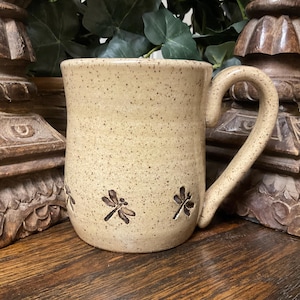 May include: A speckled beige ceramic mug with a curved handle. The mug features three dragonfly designs in brown. The mug is sitting on a wooden surface.