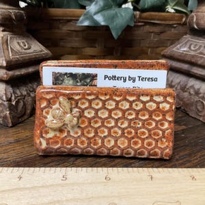 May include: A ceramic business card holder with a honeycomb pattern and a bee accent. The holder is brown and tan with the text "Pottery by Teresa" on a card inside. The holder is on a wooden surface.