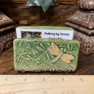May include: A rectangular, green ceramic business card holder with a dragonfly design. The holder has a raised dragonfly and leaf pattern. The text "Pottery by Teresa" is visible on a card inside the holder.