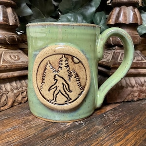 May include: A light green ceramic mug with a speckled finish and a large, circular tan medallion on the front. The medallion features a dark brown embossed illustration of a Bigfoot creature walking through a forest of pine trees under a small moon.