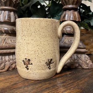 May include: A speckled beige ceramic mug with a large curved handle. Two brown frog silhouettes are etched on the side. The mug has a rustic, handmade appearance, suitable for coffee or tea.