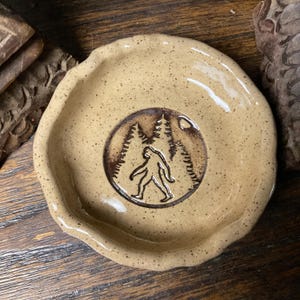 May include: A round, beige ceramic dish with a speckled finish and a wavy, uneven rim. The center of the dish features a dark brown, circular stamped design depicting a silhouette of a Bigfoot figure standing in front of a forest of pine trees.