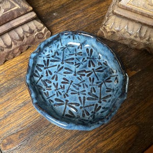 May include: A round ceramic dish with a wavy, uneven rim, featuring a glossy blue glaze and a repeating pattern of dark brown dragonfly silhouettes across the entire interior surface, creating a textured and detailed appearance.