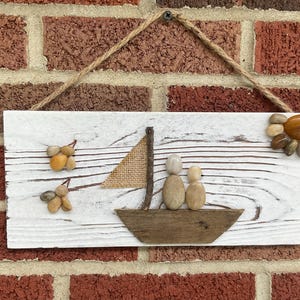 May include: A rustic wooden sign with a sailboat scene. The boat, sail, and figures are crafted from natural materials like wood and pebbles. The sign is painted white with a distressed finish and hangs from a jute rope.