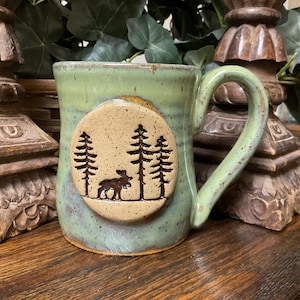 May include: A handmade ceramic mug with a green glaze and a brown handle. The mug features a circular emblem with a forest scene, including trees and a moose. The mug is designed for hot beverages.