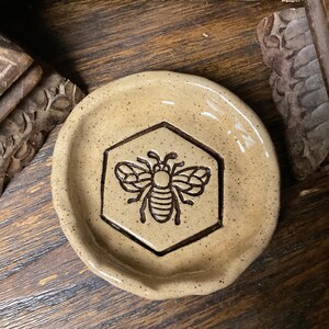 May include: A round, speckled beige ceramic dish with a slightly uneven, hand-formed rim. The center features a dark brown, embossed hexagonal outline containing a detailed, stylized illustration of a bee with wings and striped body.