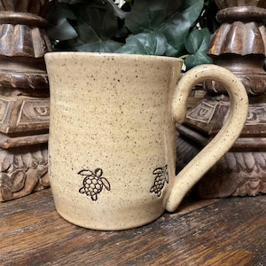 May include: A beige ceramic mug with a speckled finish and a curved handle. The exterior surface features small, dark brown stamped illustrations of sea turtles arranged in a horizontal row around the lower portion of the cylindrical body.