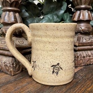 May include: A beige ceramic mug with a speckled finish and a large curved handle. The exterior features small, dark brown stamped sea turtle designs with spiral shell patterns arranged around the lower portion of the cylindrical body.