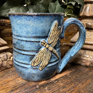 May include: A ceramic mug with a blue glazed finish and horizontal dark brown stripes. A raised, textured beige dragonfly relief is applied to the front of the mug. The piece features a sturdy, curved handle and a speckled, uneven surface texture.