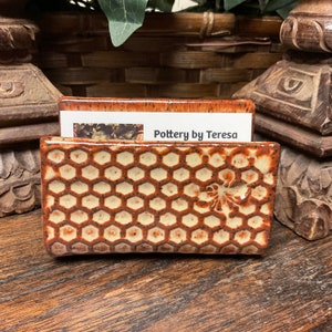 May include: A rectangular ceramic business card holder with a honeycomb pattern and a bee design. The holder is brown and cream-colored, with the text "Pottery by Teresa" on a card inside.