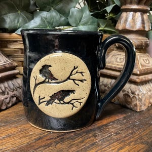 May include: A glossy black ceramic mug featuring a circular, speckled tan medallion on the front. The medallion displays a dark, etched illustration of two birds perched on bare tree branches, one above the other, with a curved handle.