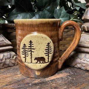 May include: A brown ceramic mug with a glossy finish and a textured surface. A circular, speckled beige medallion is attached to the front, featuring a dark brown embossed graphic of a bear walking among three tall pine trees.