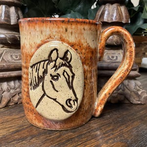 May include: A handmade ceramic mug with a horse head relief. The mug has a speckled orange and brown glaze with a curved handle. The horse head is tan with brown details. The mug is a rustic style.